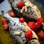 Dorade aus der Fischpfanne mit Kirschtomaten und Knoblauch auf einer Servierplatte angerichtet.