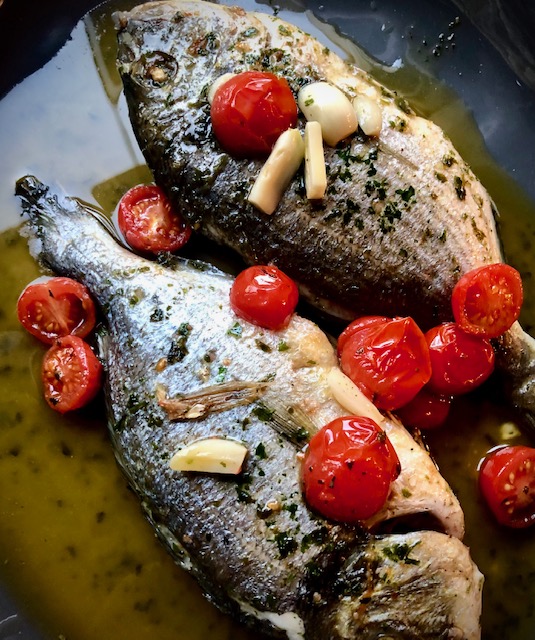 Dorade aus der Fischpfanne mit Kirschtomaten und Knoblauch auf einer Servierplatte angerichtet.