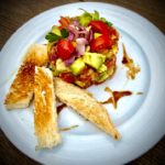 Avocado-Tomaten-Tatar auf einem weißen Teller angerichtet und ein paar Streifen Toastbrot dazu.