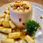Panna Cotta mit weißem Spargel und gerösteten Pinienkernen auf einem dekorativen Teller angerichtet.