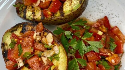 Zwei gegrillte Avocado mit Tomaten-Salsa gefüllt auf einem weißen Teller mit etwas Tomaten-Salsa und Petersilie angerichtet.