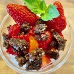 Quarkcreme mit frischen Erdbeeren und karamelisiertem Pumpernickel-Crunch in einer Glasschale serviert.