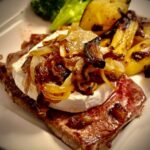 Ein Camembert-Steak mit einem ganzen runden Camembert auf dem Steak mit gerösteten Zwiebeln. Im HIntergrund zwei Röschen vom Brokkoli und angebratene Kartoffelspalten auf einem Teller angerichtet.
