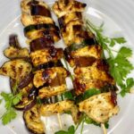 Auf der Teppanyake-Platte gegrillte Hähnchen-/Zucchini-Spieße auf einem weißen Teller angerichtet und mit frischer Petersilie garniert.