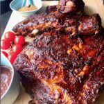 Spareribs auf einem Holzbrett mit Tomaten und BBQ-Sauce angerichtet.