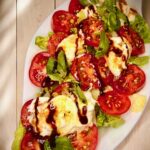 Tomatensalat mit Burrata und frischem Basilikum in einer Öl-Balsamico-Sauce auf weißem Teller angerichtet.