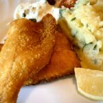 Zwei Stücke panierter Saibling mit Kartoffelsalat und Remoulade auf einem weißen Teller mit Zitrone angerichtet.