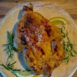 Ein ganzes leicht gebräuntes Huhn aus dem Ofen auf einem Teller mit Rosmarien und Zitrone angerichtet.