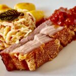Eine Scheibe Krustenbraten mit Krautsalat und Kartoffeln auf einem weißen Teller.
