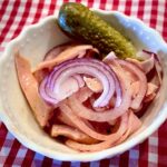 Leberkäs-Wurstsalt in einer kleinen weißen Schüssel mit einer sauren Gurke und roten Zwiebeln angerichtet.
