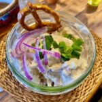 Obatzter in einem Glas angerichtet und mit Zwiebeln und Schnittlauch und einer kleinen Bezen garniert.