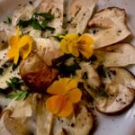 Auf einem Tisch liegt ein weißer Teller mit Steinpilz Carpaccio, dekoriert mit essbaren Blumen und Parmesan.