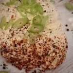 Frischkäse sebst gemacht auf einem Holzbrett mit etwas Chili und Frühlingszwiebeln kleingeschnitten angerichtet.