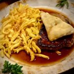 Ein klassischer schwäbischer Zwiebelrostbraten mit Spätzle und einer Maultasche und einer kräftigen dunklen Soße serviert.