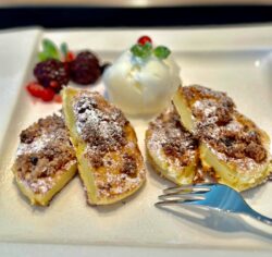 Auf einem weißen Teller liegen vier goldbraune, mit Puderzucker bestreute Ananas-Küchlein. Dazu gibt es eine weiße Eiskugel, die von glänzenden Beeren und einem Minzblatt umgeben ist. Eine silberne Gabel liegt schon bereit, um dieses süße Dessert zu kosten.