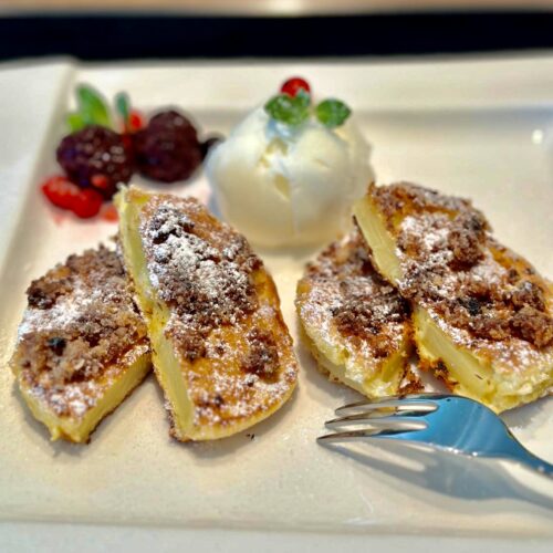 Auf einem weißen Teller liegen vier goldbraune, mit Puderzucker bestreute Ananas-Küchlein. Dazu gibt es eine weiße Eiskugel, die von glänzenden Beeren und einem Minzblatt umgeben ist. Eine silberne Gabel liegt schon bereit, um dieses süße Dessert zu kosten.