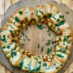 Pizza als Kranz geformt mit frischem grünem Pesto und Cheddar-Käse gefüllt auf einer grauen Schieferplatte dekoriert und mit Kräutern oben drauf.
