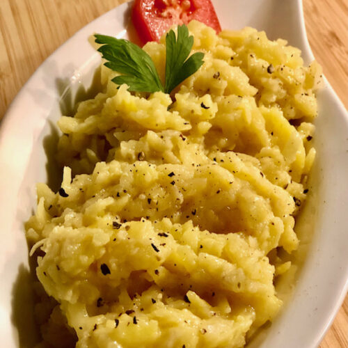 Ein geriebener Kartoffelsalat mit Petersilie und Tomate garniert auf ein einer kleinen weißen Schüssel.