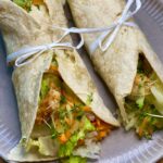 Wrap's mit gebratenen Hähnchenstreifen, Käse und Salat auf einem hellen Teller angerichtet und mit einer Schleife dekoriert.