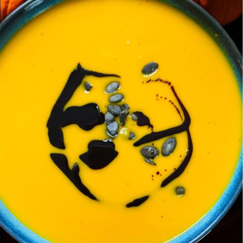 Kürbiscremesuppe in einer blauen Schale mit Kürbiskernöl und Kürbiskernen dekoriert. Im Hintergrund Kürbisse