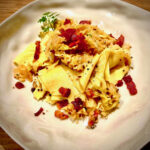 Nahaufnahme von cremigen, winterlichen Wirsing-Fleckerl (Krautfleckerl) mit rautenförmigen Nudeln, geschmortem Wirsingkraut mit Kümmel und reichlich kross ausgebratenen Speckstreifen, angerichtet auf einem hellen, eckigen Teller.