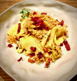 Nahaufnahme von cremigen, winterlichen Wirsing-Fleckerl (Krautfleckerl) mit rautenförmigen Nudeln, geschmortem Wirsingkraut mit Kümmel und reichlich kross ausgebratenen Speckstreifen, angerichtet auf einem hellen, eckigen Teller.