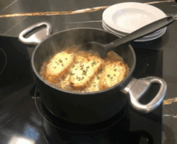 Servierfertige französische Zwiebelsuppe im Harecker Topf auf dem Herd, mit Schöpfkelle und bereitgestellten Tellern im Hintergrund.