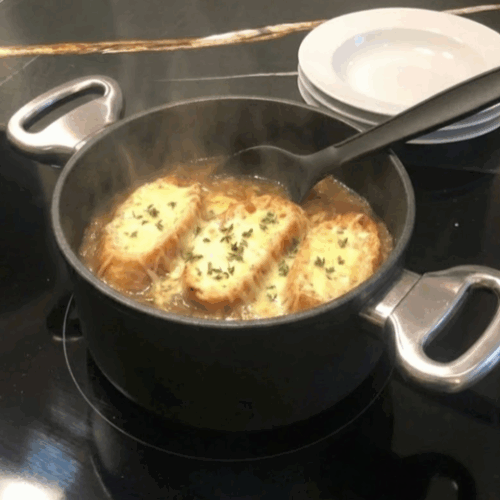 Servierfertige französische Zwiebelsuppe im Harecker Topf auf dem Herd, mit Schöpfkelle und bereitgestellten Tellern im Hintergrund.