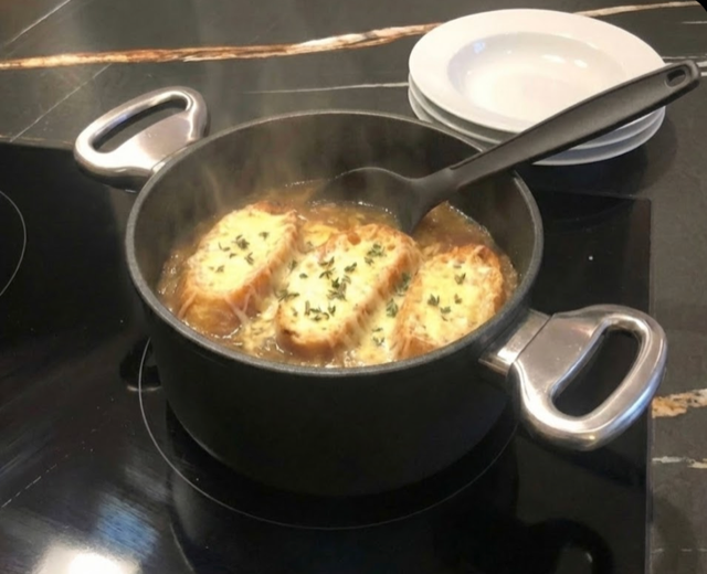 Servierfertige französische Zwiebelsuppe im Harecker Topf auf dem Herd, mit Schöpfkelle und bereitgestellten Tellern im Hintergrund.