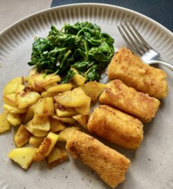 Fischstäbchen aus frischem Kabeljaufilet mit Bratkartoffeln und frischem Spinat aus der Pfanne. Auf einem hellen Teller angerichtet und noch etwas Pfeffer darüber gegeben.