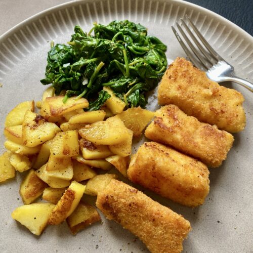 Fischstäbchen aus frischem Kabeljaufilet mit Bratkartoffeln und frischem Spinat aus der Pfanne. Auf einem hellen Teller angerichtet und noch etwas Pfeffer darüber gegeben.