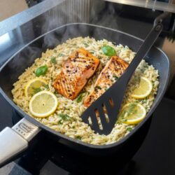 Gebratenes Lachsfilet auf Orzo-Pasta (Kritharaki) mit Zitronenscheiben und frischen Kräutern in einer quadratischen, schwarzen Pfanne auf einem Induktionsherd.