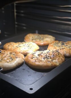 Sechs goldbraune, mit schwarzem Sesam bestreute Fladenbrote backen auf einem schwarzen Backblech in einem Ofen.