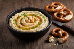 Eine Schale cremiger bayerischer Hummus mit Kichererbsen, Olivenöl und Schnittlauch, serviert mit bayerischen Laugenbrezeln auf einem dunklen Holztisch.