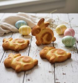 Vier goldbraun gebackene Osterhasen aus Rührteig auf einem rustikalen weißen Holztisch, dekoriert mit bunten Ostereiern und Leinentuch.