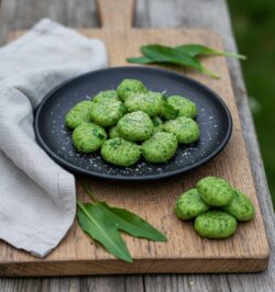 Grüne Bärlauch-Gnocchi mit Parmesan bestreut auf einem schwarzen Teller, serviert auf einem Holzbrett neben frischen Bärlauchblättern.