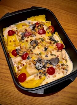 Gefüllte Ravioli in cremiger Sauce mit schwarzen Trüffelscheiben, Kirschtomaten und gerösteten Pinienkernen in einer rechteckigen schwarzen Auflaufform von Pfannen Harecker.