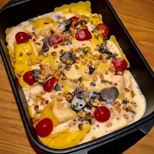 Gefüllte Ravioli in cremiger Sauce mit schwarzen Trüffelscheiben, Kirschtomaten und gerösteten Pinienkernen in einer rechteckigen schwarzen Auflaufform von Pfannen Harecker.