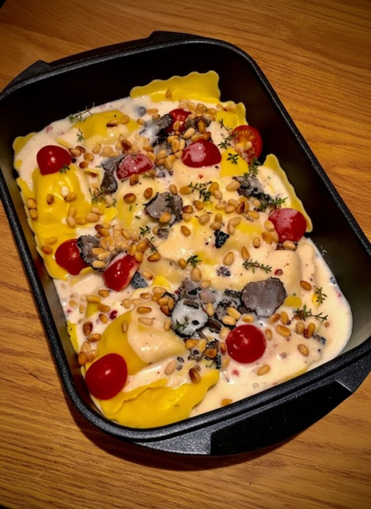 Ravioli-Auflauf mit Trüffel in cremiger Sauce mit schwarzen Trüffelscheiben, Kirschtomaten und gerösteten Pinienkernen in einer rechteckigen schwarzen Auflaufform von Pfannen Harecker.