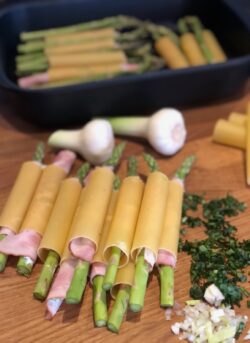 Grüner Spargel mit Schinken umwickelt in Cannelloni-Pasta auf einem Holzbrett neben einer dunklen Auflaufform und frischen Kräutern.