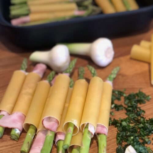 Grüner Spargel mit Schinken umwickelt in Cannelloni-Pasta auf einem Holzbrett neben einer dunklen Auflaufform und frischen Kräutern.
