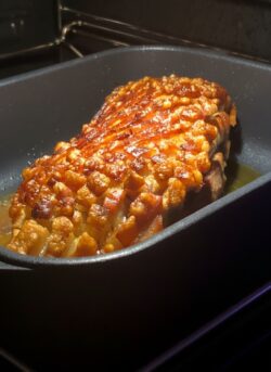 Frisch zubereiteter Schweinebraten mit goldbraun gebackener, knuspriger Kruste in einem dunklen Bräter im Backofen.